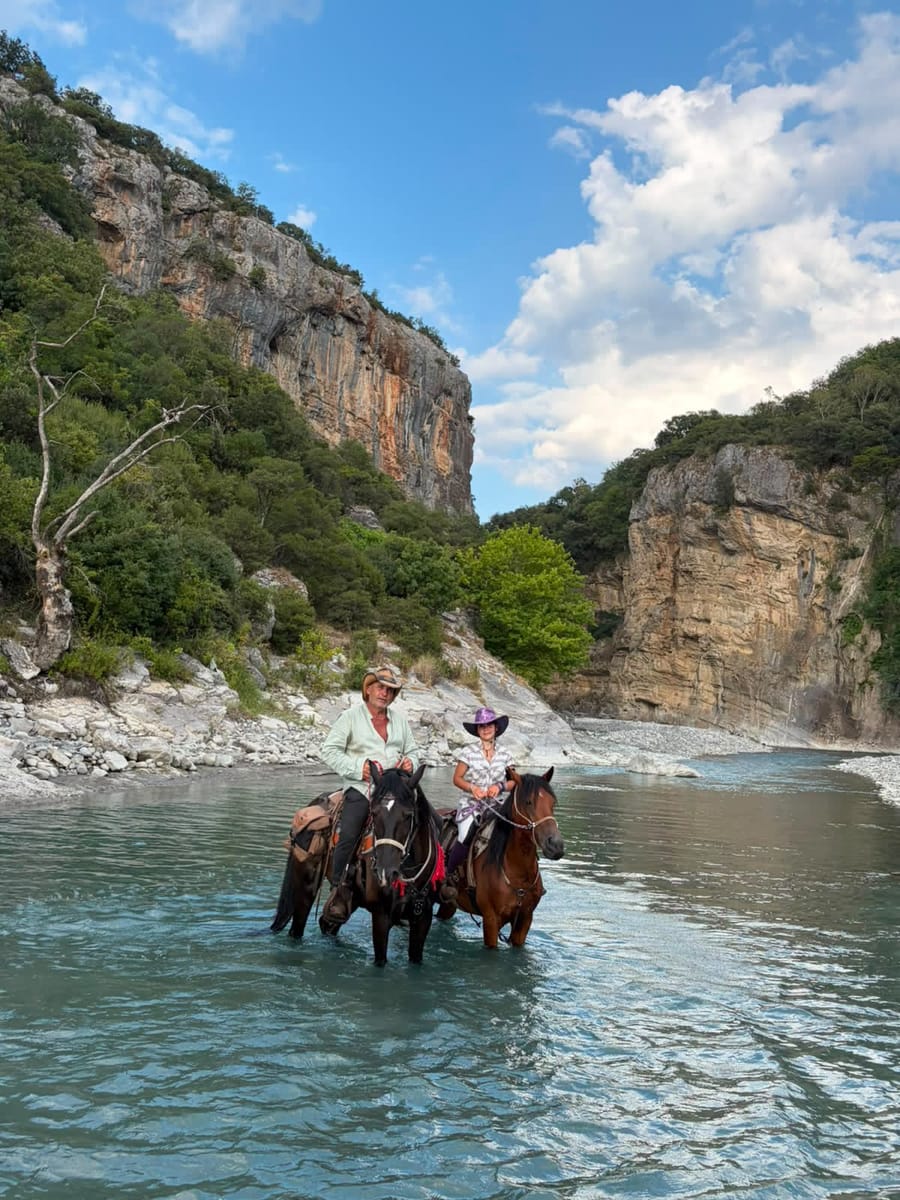 Two riders on horseback in a river below limestone cliffs