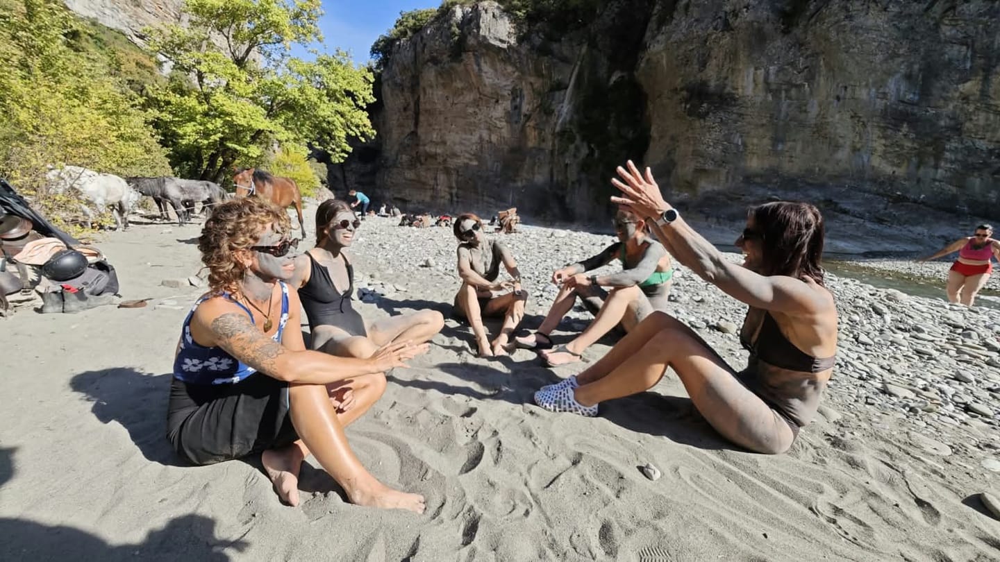 Riders relaxing on a sandy riverbank below cliffs